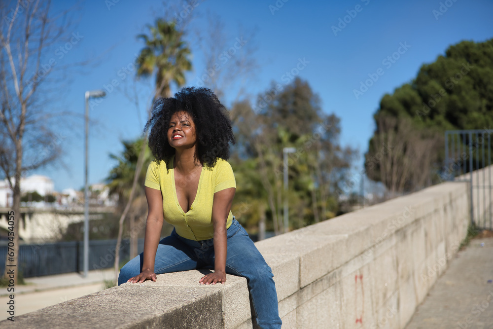 Young, beautiful, black woman with afro hair, wearing yellow t-shirt and jeans, sitting astride a stone wall, smiling, happy and sexy. Concept laughter, happiness, sensuality.