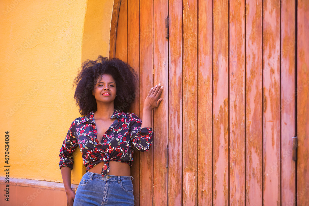 Fototapeta premium Young woman, beautiful and black with afro hair, with a floral shirt leaning on a wooden board door, quiet, happy and smiling. Concept beauty, tranquility, happiness.