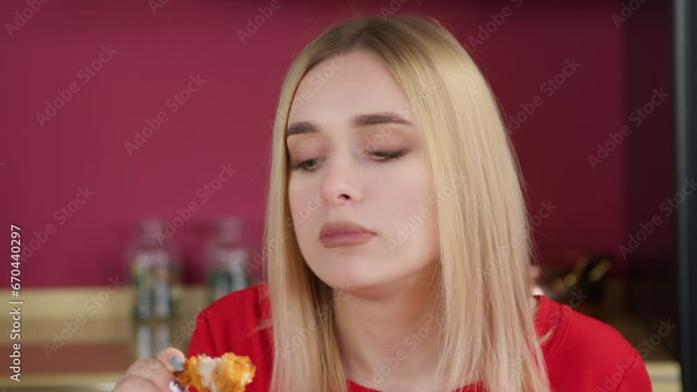 Close up. A young blonde woman on the background of a red black room of the kitchen eats a chicken wing while savoring it. Concept of eating fast food. Close up. A young blonde woman on the background of a red black room of the kitchen eats a chicken wing while savoring it. Concept of eating fast food.