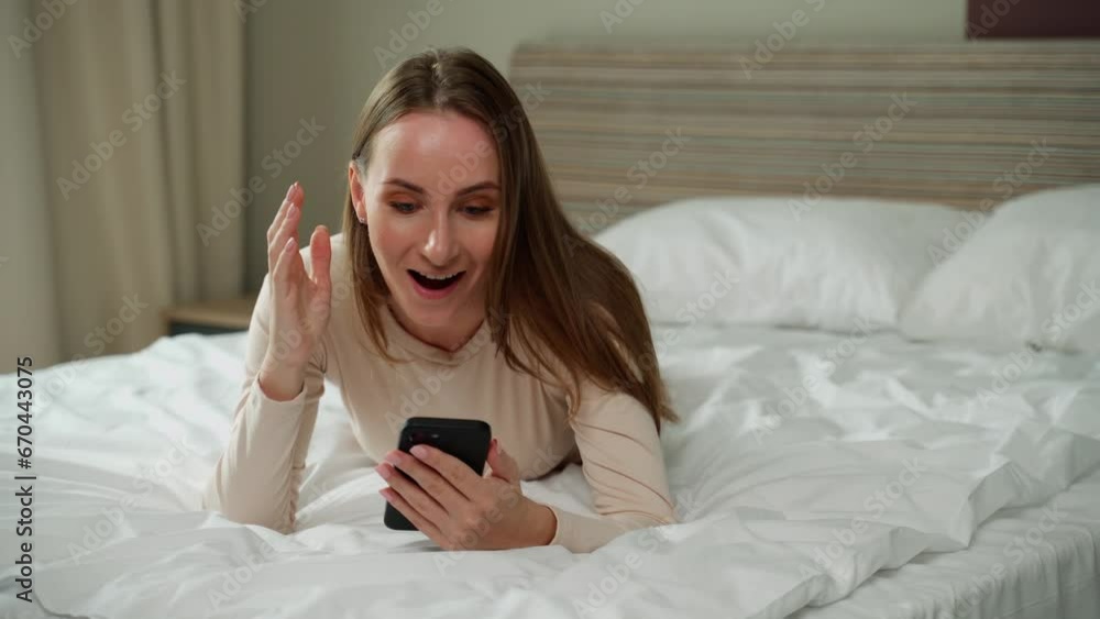 The excited winner checks the online phone in search of a prize while lying at home on the bed. Victorious and triumphant. An excited girl in bed, looking at her phone with an astonished expression on The excited winner checks the online phone in search of a prize while lying at home on the bed. Victorious and triumphant. An excited girl in bed, looking at her phone with an astonished expression on