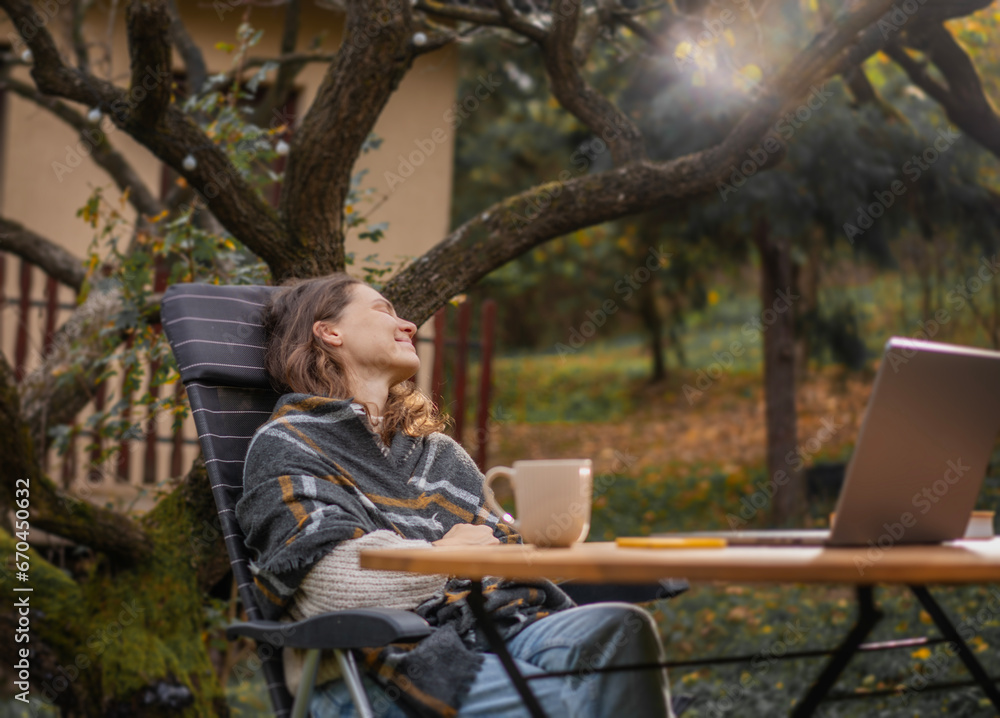 A young woman sitting in the autumn garden at a table with a laptop enjoying the fresh air and nature A young woman sitting in the autumn garden at a table with a laptop enjoying the fresh air and nature