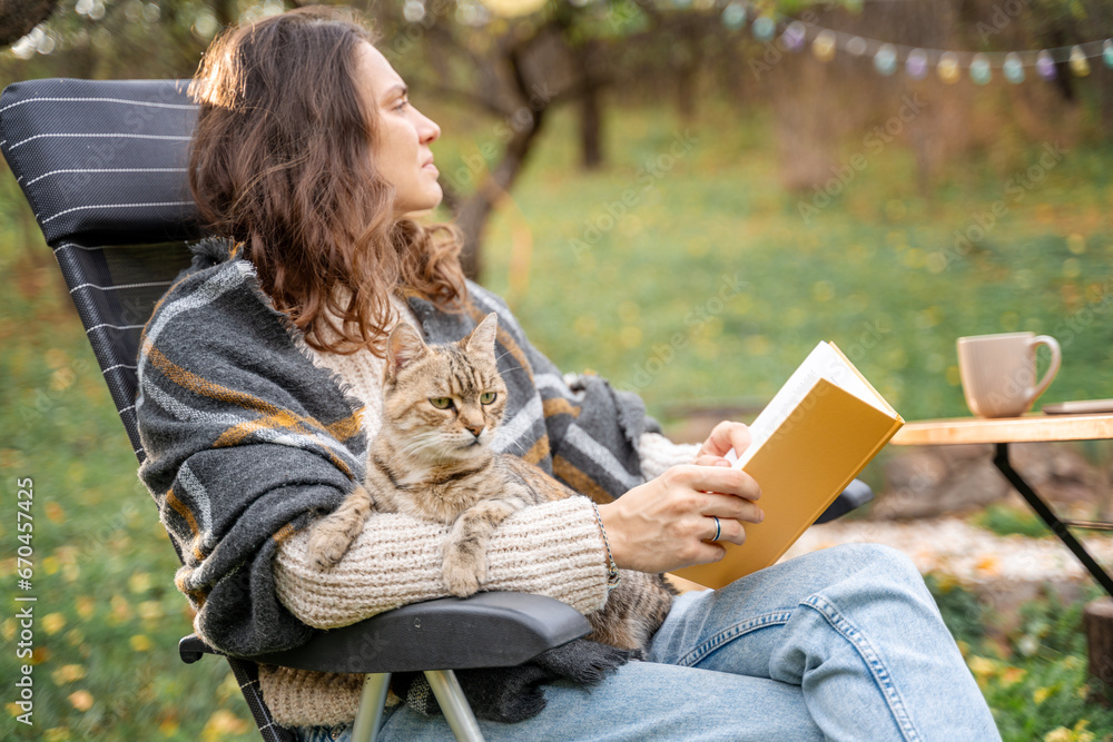 Obraz premium Young woman sitting in the autumn garden with a pet gray cat in her hands reading a book