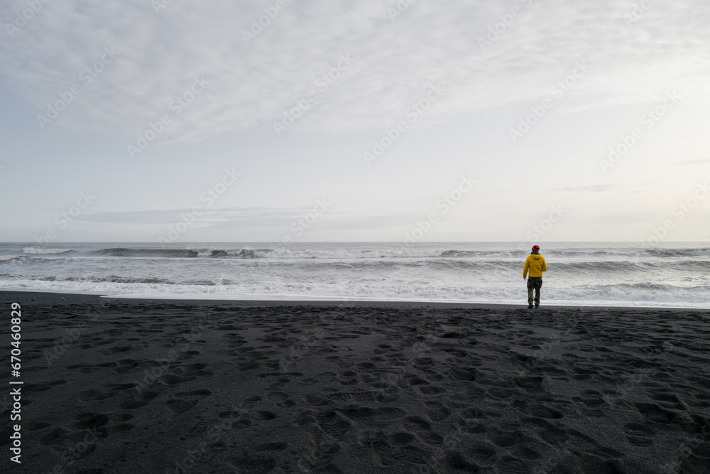 Obraz premium Man in a yellow jacket standing on the black sand beach overlooking the horizon
