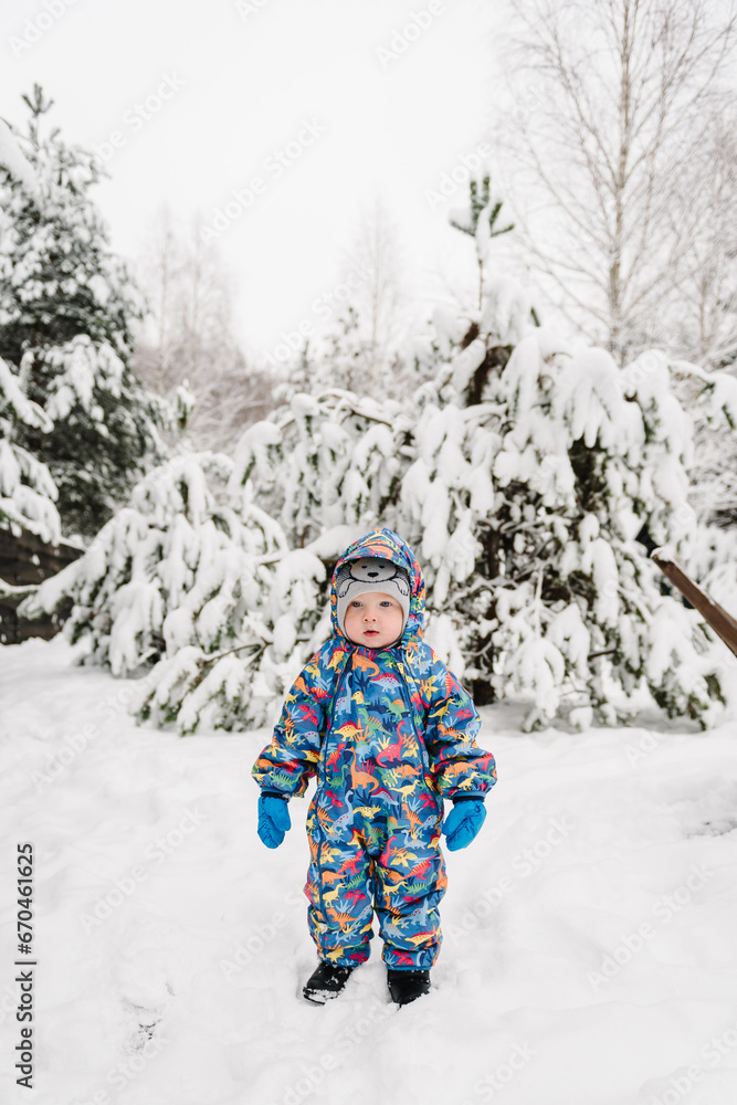 Little child in blue winter clothes having fun. Kid wearing warm hat walking in winter park in holidays. Toddler in mountain country in snowy forest. Baby boy playing snow among snowdrifts closeup.