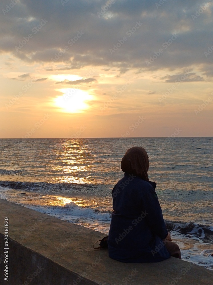 Woman sitting on a wall watching the sunset over the sea in summer