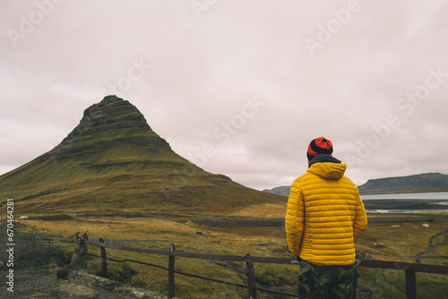 Φωτογραφία View from behind on man in a yellow jacket overlooking Kirkjufell mountain in Ic