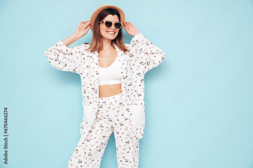Young beautiful smiling female in trendy summer costume clothes. Carefree woman posing near blue wall in studio. Positive model having fun indoors. Cheerful and happy. In hat, sunglasses