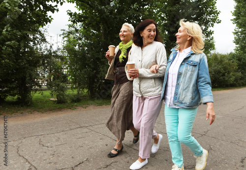 Three elderly ladies in casual clothes are walking in the park. Coffee and socializing, friendship in retirement.