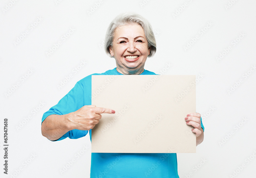 A happy and charming elderly woman holds a blank sheet of paper in her hands and points at it with her finger. A happy and charming elderly woman holds a blank sheet of paper in her hands and points at it with her finger.