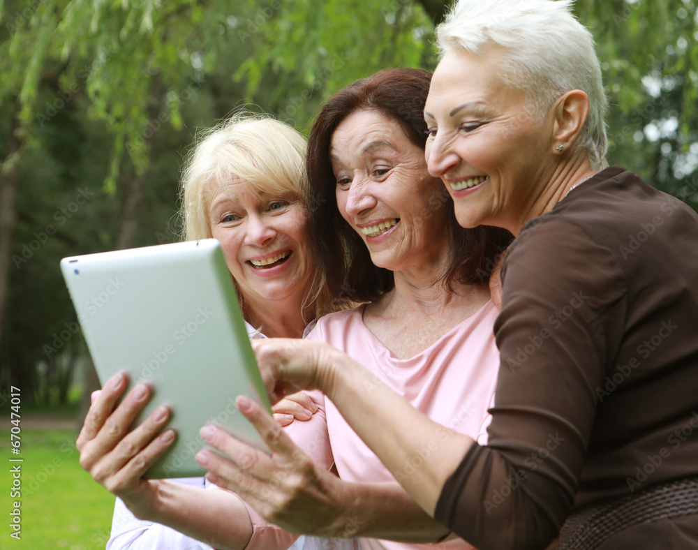 Three old women are smiling and looking photos at the at screen of the tablet pc in the park on a summer day Three old women are smiling and looking photos at the at screen of the tablet pc in the park on a summer day