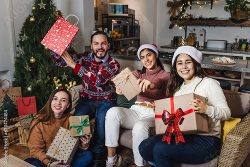 portrait of Hispanic friends holding gifts and sitting near Christmas tree at home in Mexico. Holidays and celebration concept in Latin America