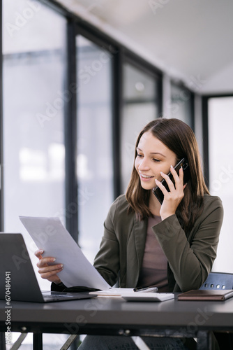 businesswoman talking mobile phone and working on laptop and papers on table in her office.