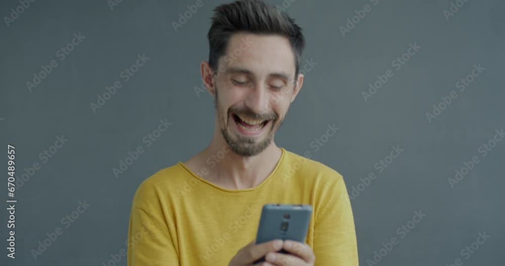 Joyful young man texting with smartphone and laughing standing on grey color background. Online communication and mobile application concept. Joyful young man texting with smartphone and laughing standing on grey color background. Online communication and mobile application concept.