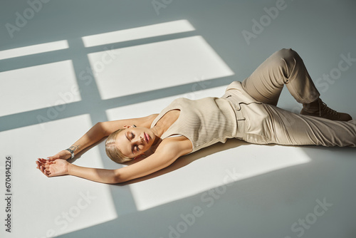 young sensual woman in beige outfit lying down with closed eyes in sunshine on grey backdrop