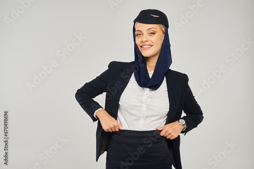 smiling arabian airlines stewardess in uniform and headscarf posing with hands on waist on grey