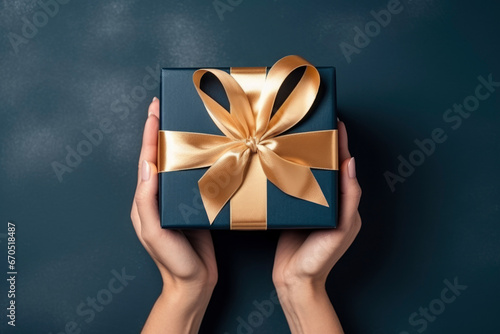 overhead view, womans hands holding a luxury gift box with gold large satin bow against a dark blue background. Close up. New Year present. photo