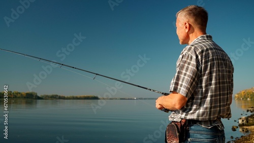 Medium close-up of an adult man in jeans and a shirt fishing on the banks of a picturesque river. A mature man with a short haircut and a little gray in his hair catches fish with a spinning rod