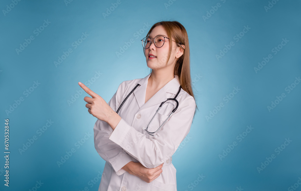 Young asian doctor wear medical uniform lab coat and stethoscope on blue background. Smiling female doctor pointing side to empty copy space for healthcare product or show banner rules safety. Young asian doctor wear medical uniform lab coat and stethoscope on blue background. Smiling female doctor pointing side to empty copy space for healthcare product or show banner rules safety.
