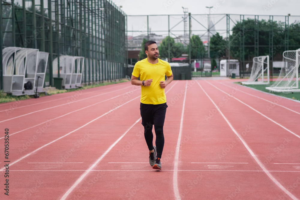 Sportive guy in tracksuit running along red track with white separating lines by soccer field on large sports ground in urban park for health and body development Sportive guy in tracksuit running along red track with white separating lines by soccer field on large sports ground in urban park for health and body development