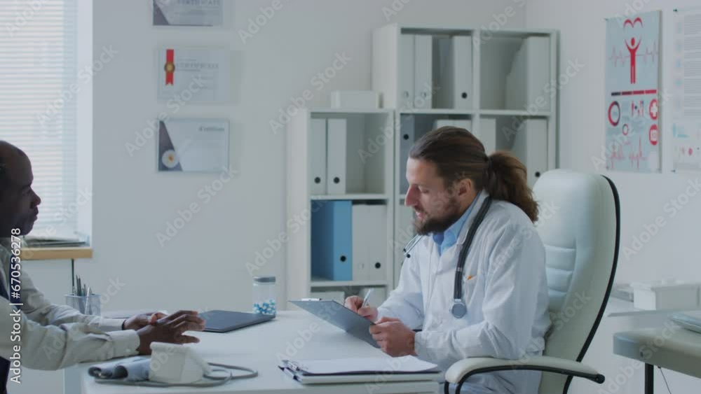 Medium pan shot of Caucasian physician talking to black male patient and writing down complaints about lungpain and cough into medical record during appointment in clinic Medium pan shot of Caucasian physician talking to black male patient and writing down complaints about lungpain and cough into medical record during appointment in clinic