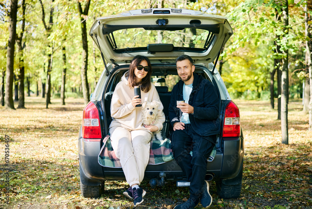 Couple with their dog relaxing in forest sitting in car trunk - obrazy, fototapety, plakaty 