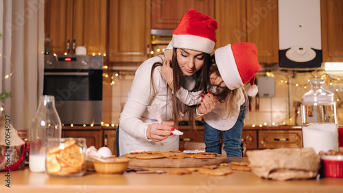Adorable little daughter with her mom at kitchen during Christmas holidays. Decorative fit tree at the table. Girls make gingerbread