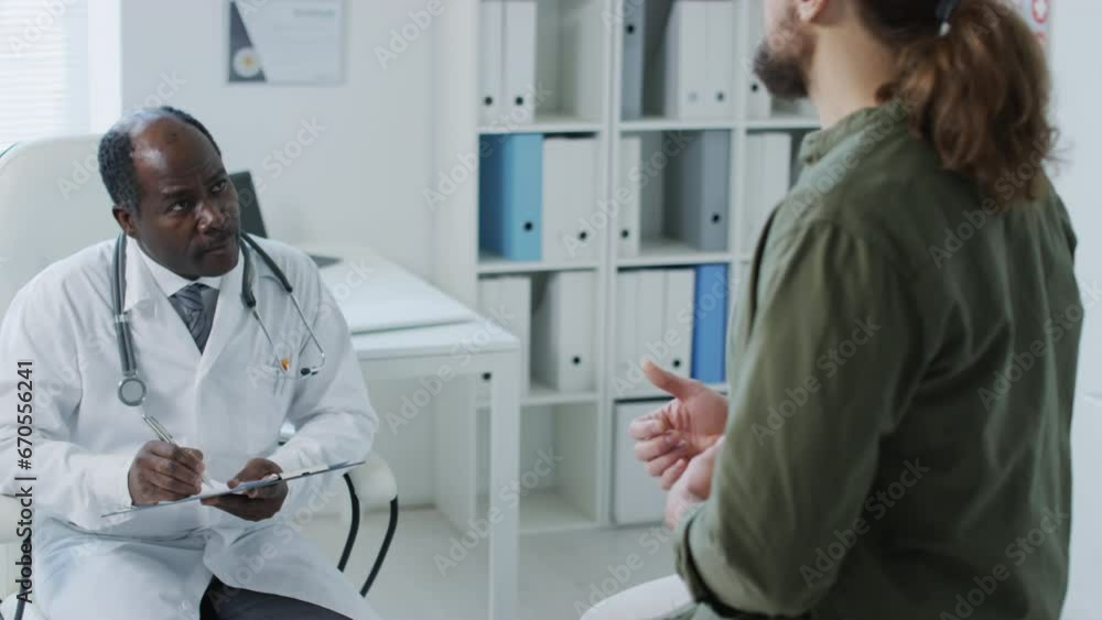 Rear view of patient speaking about complaints of pain with black male doctor writing information down in medical history during appointment in hospital Rear view of patient speaking about complaints of pain with black male doctor writing information down in medical history during appointment in hospital