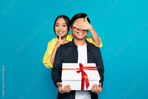 Smiling millennial asian woman in casual closed eyes to man, hold gift box with present photo