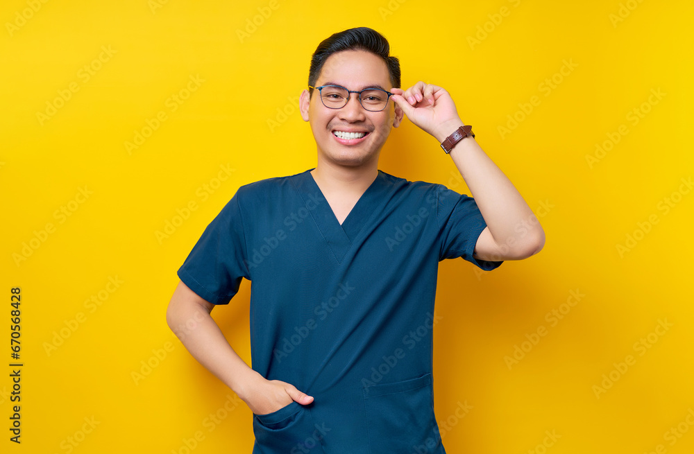 Handsome young Asian man doctor or nurse wearing a blue uniform and glasses confident looking at the camera with smiling friendly isolated on yellow background. Healthcare medicine concept Handsome young Asian man doctor or nurse wearing a blue uniform and glasses confident looking at the camera with smiling friendly isolated on yellow background. Healthcare medicine concept