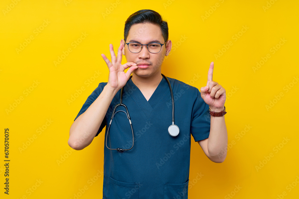 Serious young Asian man doctor or nurse wearing a blue uniform and glasses makes a zipping mouth gesture, promising to keep secret, pointing up isolated on yellow background Serious young Asian man doctor or nurse wearing a blue uniform and glasses makes a zipping mouth gesture, promising to keep secret, pointing up isolated on yellow background