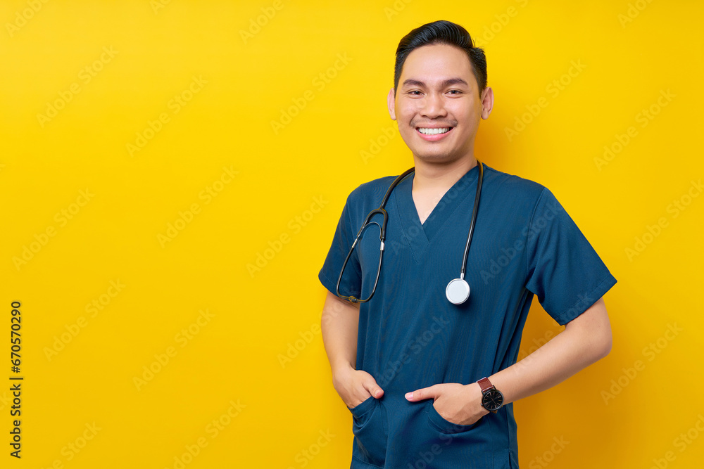 Professional young Asian man doctor or nurse wearing a blue uniform and stethoscope standing confidently while smiling friendly at camera, hands in pockets isolated on yellow background Professional young Asian man doctor or nurse wearing a blue uniform and stethoscope standing confidently while smiling friendly at camera, hands in pockets isolated on yellow background