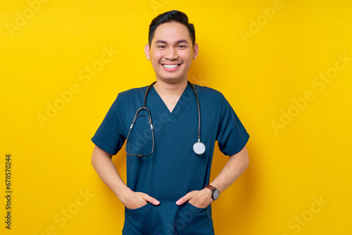 Professional young Asian man doctor or nurse wearing a blue uniform and stethoscope standing confidently while smiling friendly at camera, hands in pockets isolated on yellow background