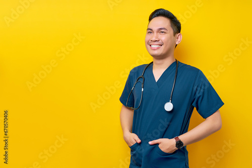 Professional young Asian man doctor or nurse wearing a blue uniform and stethoscope standing confidently while smiling friendly aside, hands in pockets isolated on yellow background photo