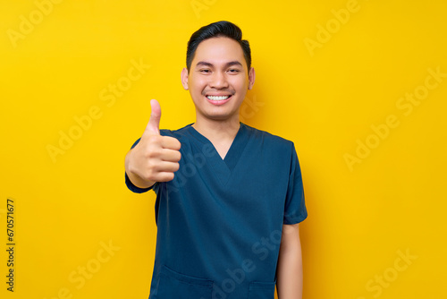 Professional young Asian male doctor or nurse wearing a blue uniform showing thumb up or like, looking at camera with smile isolated on yellow background. Healthcare medicine concept