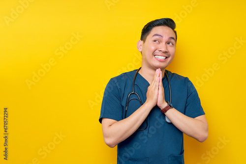 Hopeful professional young Asian male doctor or nurse wearing a blue uniform begging for help, pleading to God isolated on yellow background. Healthcare medicine concept photo