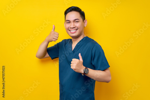Professional young Asian male doctor or nurse wearing a blue uniform showing thumb up or like, looking at camera with smile isolated on yellow background. Healthcare medicine concept