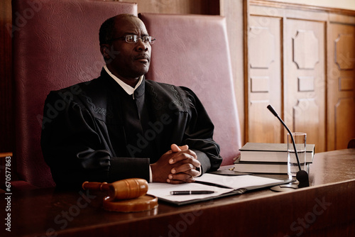 Serious male impartial judge in uniform sitting by desk in front of microphone and keeping his hands clasped during trial session
