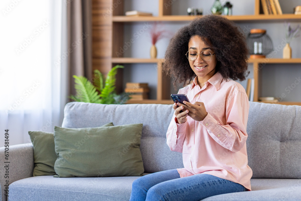 Young beautiful woman with curly hair is using the phone, typing messages and browsing online internet pages, sitting at home on the sofa in the living room of the house. Young beautiful woman with curly hair is using the phone, typing messages and browsing online internet pages, sitting at home on the sofa in the living room of the house.