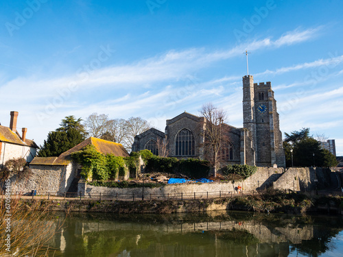 In foreground, the River Medway flows past All Saints Church, a paved riverside walkway and period stone houses at Maidstone, Kent. Bright blue sky, light clouds.