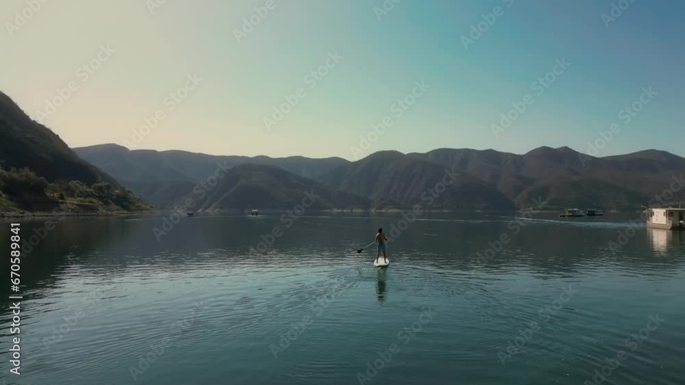 Aerial drone flying over a lake, man doing stand up paddle, with mountains at sunrise. SUP. Beautifull dreamy landscape view. Cosatline with boats. Paceful landscape. Cabra Corral, Salta, Argentina.