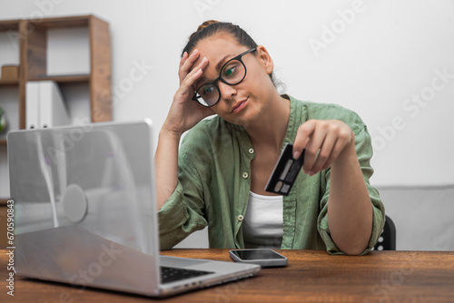 A stressed woman gazes at her laptop screen, visibly upset by her dwindling credit card balance while managing bills and online shopping. 