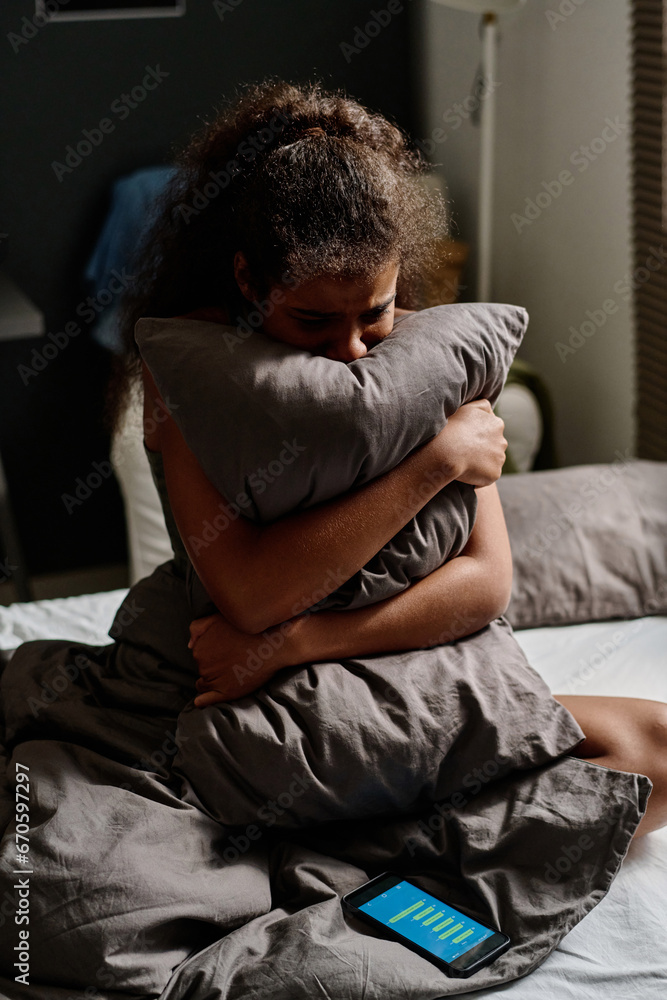 Miserable teenage girl crying in pillow while sitting on double bed with smartphone at night and suffering from cyberbullying messages Miserable teenage girl crying in pillow while sitting on double bed with smartphone at night and suffering from cyberbullying messages