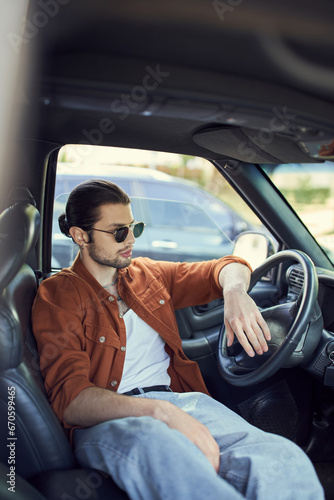 charming man with ponytail and sunglasses relaxing behind steering wheel and looking away, fashion © LIGHTFIELD STUDIOS