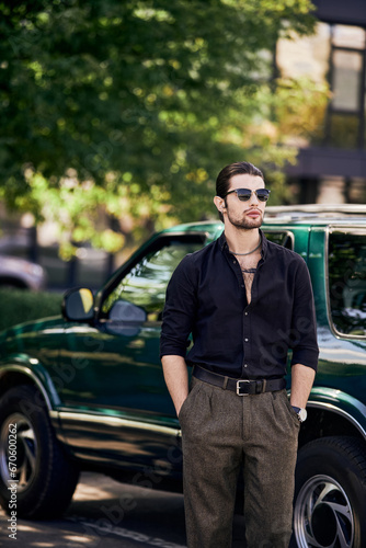 vertical shot of charming man in elegant black attire posing next to car with hands in pockets
