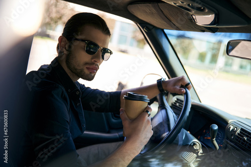 appealing man in black attire holding steering wheel with coffee cup in hand and looking at camera © LIGHTFIELD STUDIOS