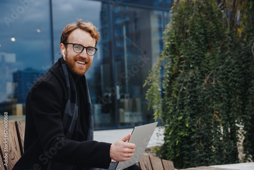 Successful businessman on the background of the business center. Red-haired young guy, in a stylish coat, wireless headphones with a laptop.