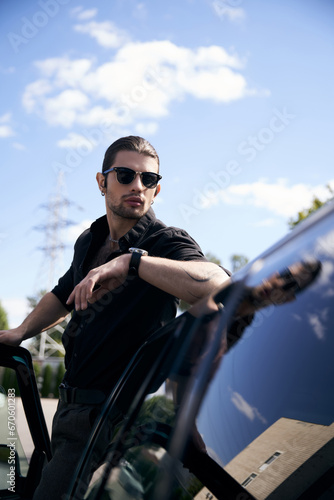handsome young male model with sunglasses and tattoo posing outdoor with arm on his car, sexy driver