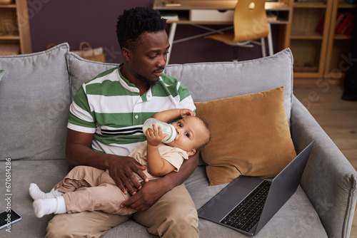 Portrait of young Black father bottle feeding cute baby boy while working from home with laptop photo