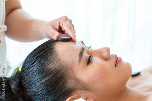 Close up therapist hand palcing black spa stone on customer forehead laying down on bed; Holistic Health and Well-Being Healing Hand with Hot Stone