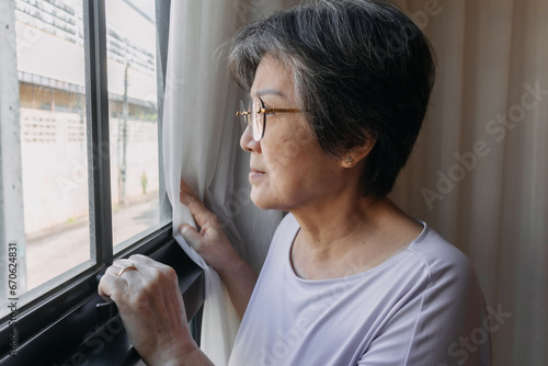 Asian old woman sitting on chair and opening white curtain while looking out the window, missing and waiting family and thinking something sad, living alone. photo